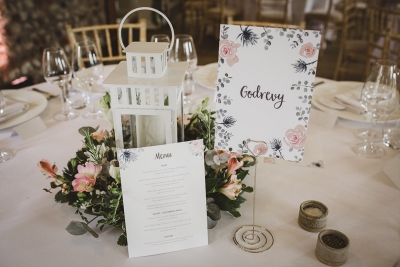A wedding reception table setup with a white lantern, floral centerpiece, and a menu card with a floral name sign.