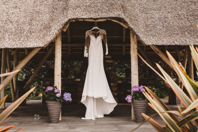 A long-sleeved white wedding dress hanging under a thatched roof, surrounded by potted plants.