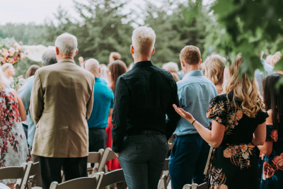 A group of wedding guests standing outdoors, dressed in formal and semi-formal attire, facing the ceremony.