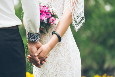 A bride in a lace wedding dress holding hands with her partner while carrying a bouquet of pink flowers.