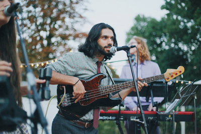 A musician playing a bass guitar and singing into a microphone during an outdoor event, with a band performing in the background.