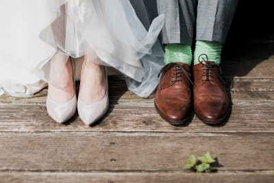 A bride in glittery heels and a groom in brown leather shoes with green socks, standing on a wooden surface.