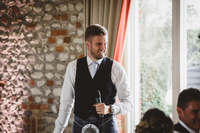 A best man in a navy waistcoat and tie holding a microphone, smiling while delivering a speech in a rustic venue.