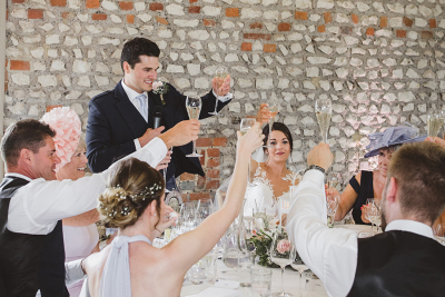 Groom gives wedding speech while guests, including the bride, raise champagne glasses for a toast in a rustic venue.