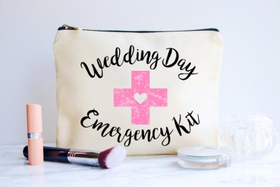 A beige makeup pouch with "Wedding Day Emergency Kit" written in black cursive and a pink cross design, surrounded by beauty products.