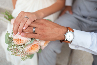 A close-up of newlyweds' hands, displaying wedding rings over a bouquet of peach roses and greenery.
