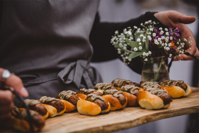 A tray of gourmet sausages on mini buns served by a waiter, with a small floral arrangement on the side.