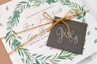 A wedding invitation featuring a botanical wreath design, tied with twine and a dark green RSVP card.