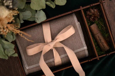 A neatly wrapped brown blanket with a beige ribbon inside a wooden gift box, surrounded by greenery and decorative elements.