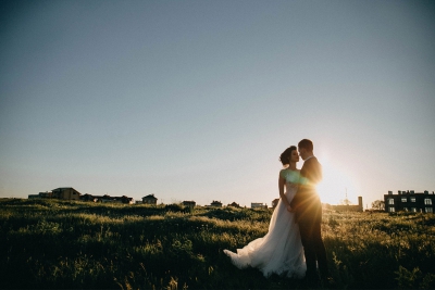 A newlywed couple standing in a grassy field at sunset, gazing at each other with silhouettes against the golden light.
