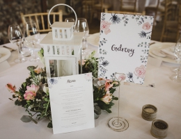 A wedding reception table setup with a white lantern, floral centerpiece, and a menu card with a floral name sign.