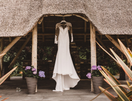 A long-sleeved white wedding dress hanging under a thatched roof, surrounded by potted plants.