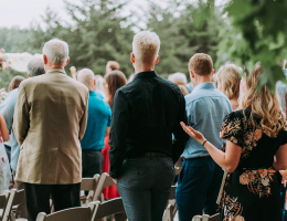 A group of wedding guests standing outdoors, dressed in formal and semi-formal attire, facing the ceremony.