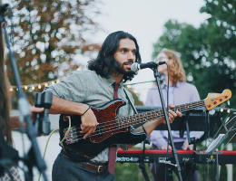 A musician playing a bass guitar and singing into a microphone during an outdoor event, with a band performing in the background.