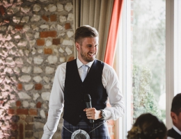 A best man in a navy waistcoat and tie holding a microphone, smiling while delivering a speech in a rustic venue.