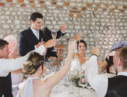 Groom gives wedding speech while guests, including the bride, raise champagne glasses for a toast in a rustic venue.