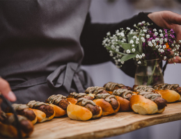 A tray of gourmet sausages on mini buns served by a waiter, with a small floral arrangement on the side.