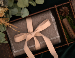 A neatly wrapped brown blanket with a beige ribbon inside a wooden gift box, surrounded by greenery and decorative elements.