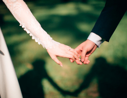 A bride and groom holding hands in soft sunlight, their shadows forming a heart shape on the grass.