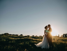 A newlywed couple standing in a grassy field at sunset, gazing at each other with silhouettes against the golden light.