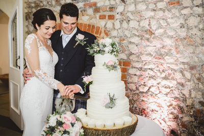 A newlywed couple smiling as they cut a white tiered wedding cake decorated with flowers in a rustic venue.