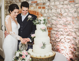 A newlywed couple smiling as they cut a white tiered wedding cake decorated with flowers in a rustic venue.
