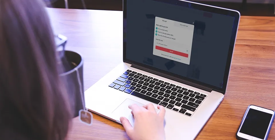 A person using a laptop with an open gift selection screen, with a phone and a tea mug on the wooden table.