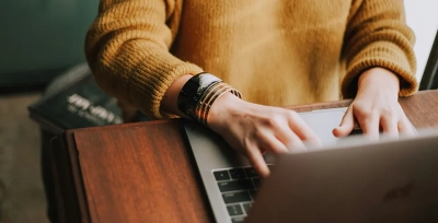 A person in a mustard sweater with gold bangles typing on a laptop at a wooden desk.
