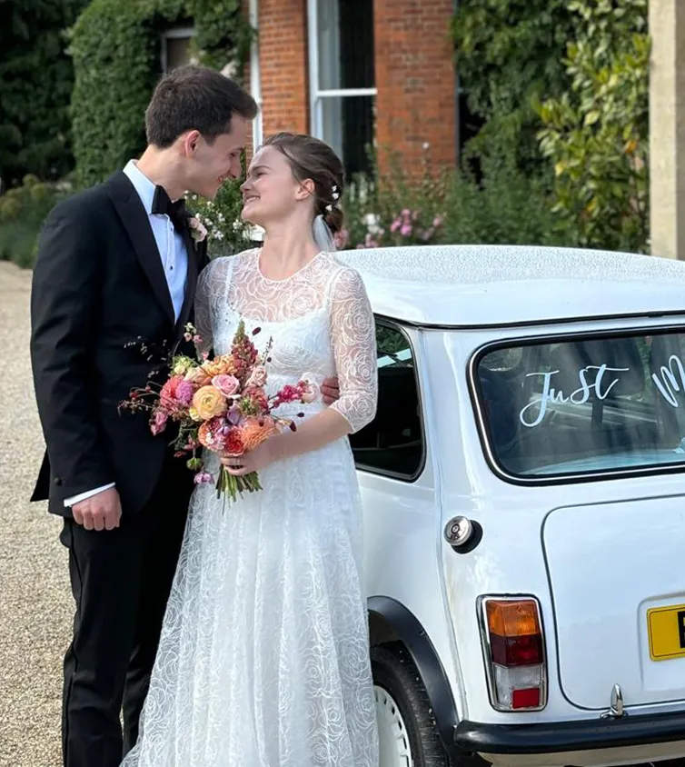 Newlyweds in formal attire stand by a white car with 'Just Married' on the window, in front of an ivy-covered brick mansion.
